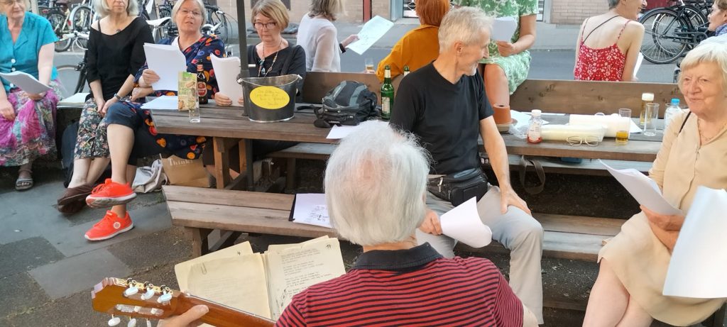 Nachbarn sitzen an Bänken auf der Straße, singen aus Liedblättern, im Vordergrund spielt jemand Gitarre.