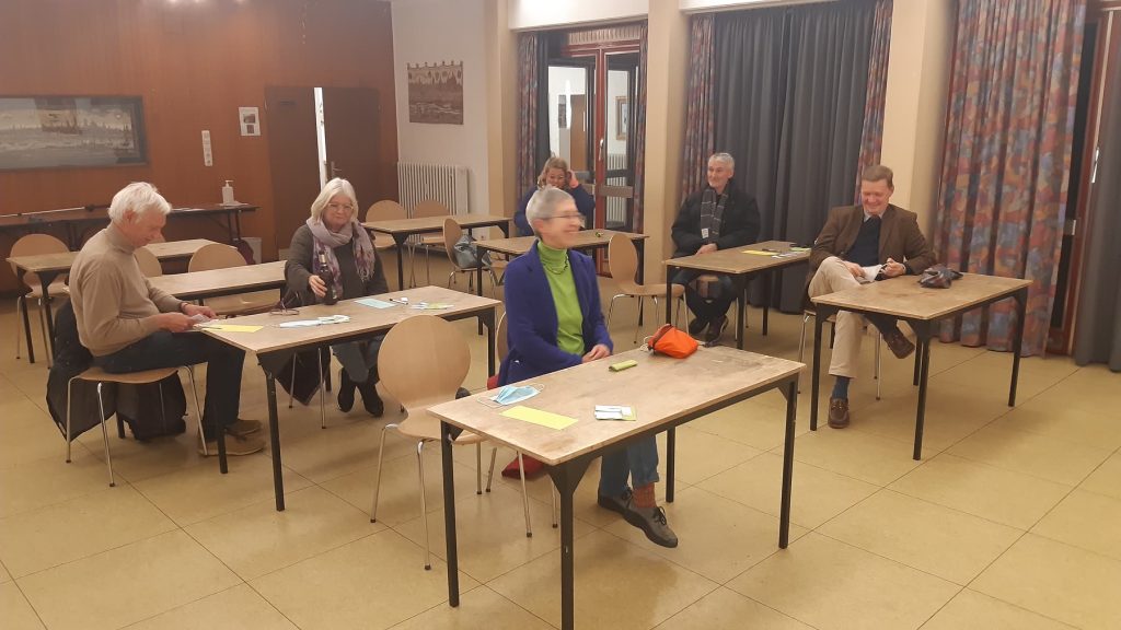 Mehrere Personen sitzen mit Abstand an einzelnen Tischen in einem Gemeinschaftsraum.